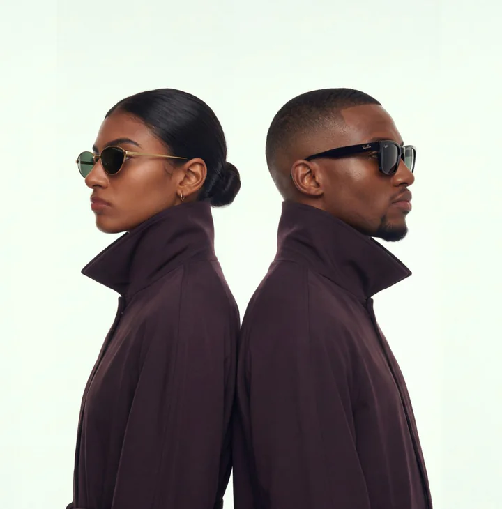 A woman and a man standing back to back in burgundy coats with high collars, seen in profile: she in thin gold-framed sunglasses, he in black square sunglasses